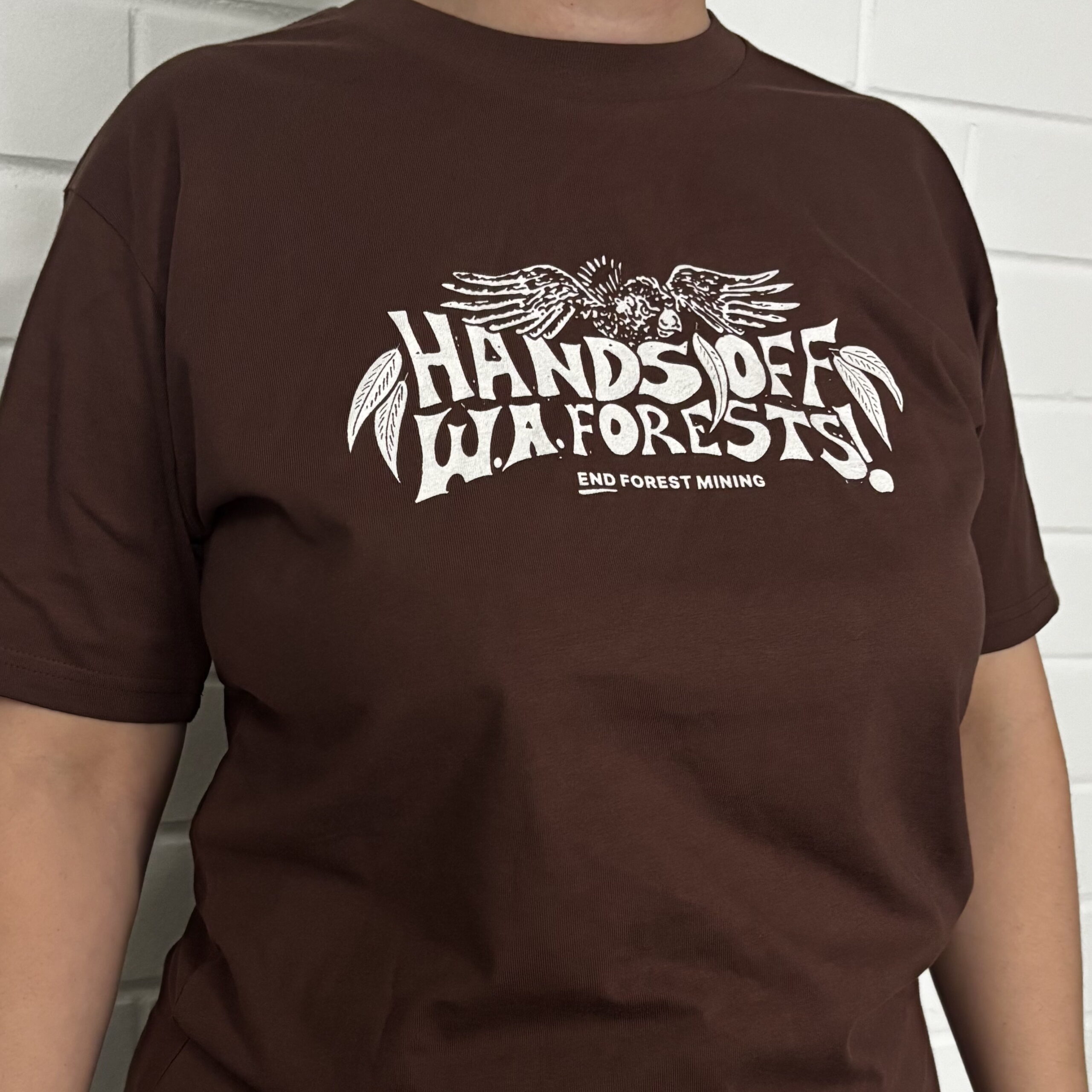 Jess wearing a brown hands off wa forests tee featuring a cockatoo and jarrah leaves.